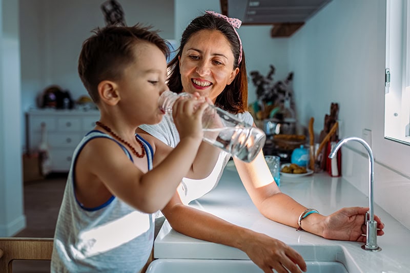 A woman smiling warmly as she watches a young boy drink water from a clear reusable bottle in a bright kitchen.